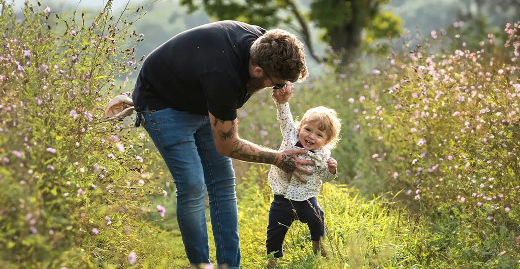 Alleinerziehender Vater spielt mit seinem kleinen Kind auf einem blühenden Weg in der Natur trotz der vielen Herausforderungen im Familienalltag.