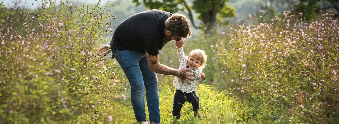 Alleinerziehender Vater spielt mit seinem kleinen Kind auf einem blühenden Weg in der Natur trotz der vielen Herausforderungen im Familienalltag.